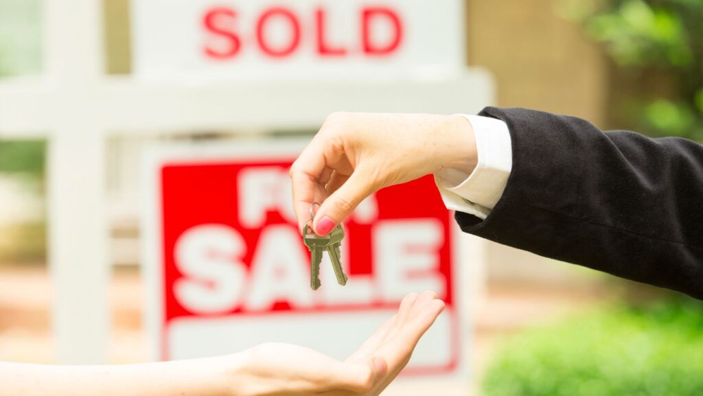 A person passing a set of house keys to another individual, against the backdrop of a "for sale" sign proudly displaying the word "sold".